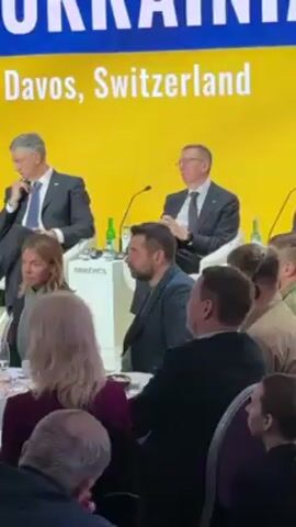 The video shows a man with a beard, wearing a blue blazer over a green shirt, sitting among other attendees at a conference or meeting. The scene is well-lit, with a bright yellow backdrop displaying the words "Davos, Switzerland." There are water bottles and microphones on the table in front of the people. The man in the blazer is looking to his right, appearing to be listening to a speaker. The atmosphere suggests a formal, professional setting.