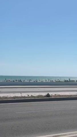 The media file depicts a sunny day at a crowded beach. The scene is viewed from across a road, with the ocean and beach stretching into the distance. The sky is a clear, bright blue, indicating good weather.

**Subjects:**
*   **People:** Numerous people are visible on the beach and in the water. They appear to be enjoying a recreational day, with many swimming, sunbathing, and walking.
*   **Objects:** Several cars are parked along the roadside and driving on the road in the foreground. There are also lamp posts, and a parking sign with a blue "P" on it is visible on the beach promenade. Umbrellas and possibly beach chairs are set up on the sand.
*   **Scene/Setting:** The setting is a beach area, likely a popular tourist destination or local recreational spot. The presence of the road and promenade suggests an urban or developed coastal area. The ocean appears calm.

**Activity or Event:**
The primary activity is a day at the beach. People are engaged in typical beach activities such as swimming, relaxing, and socializing. The presence of many cars and people suggests a busy day, possibly a weekend or holiday.

**Notable Details:**
*   **Time of Day/Weather:** The bright blue sky and the shadows cast suggest it is daytime, likely mid-morning to afternoon, under clear, sunny weather conditions.
*   **Location Cues:** The mention of Ömirzaq, Kazakhstan, places the scene in this specific region.
*   **Emotions:** While not explicitly stated, the general atmosphere appears relaxed and leisurely, indicative of a pleasant day out.

**Visible Text:**
A blue parking sign with a white "P" is visible on the promenade.