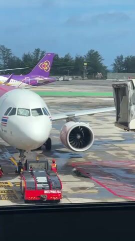 The video shows an airport tarmac through a large window. In the foreground, a red and white AirAsia airplane is parked at a gate, with its front landing gear attached to a red tug vehicle. Ground crew in orange vests are visible near the airplane. To the left, another tug vehicle is positioned. In the background, a purple and pink Thai Airways airplane is parked. The sky is partly cloudy. The scene suggests preparations for or after a flight.