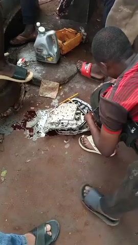 The scene shows several people gathered outdoors around what appears to be a disassembled or damaged automotive component, possibly part of an automatic transmission. The ground is dirt or packed soil, stained with what looks like oil or fluid. A container of Mobil Super oil is visible next to the object. The people are wearing casual clothing, including sandals and flip-flops. They are working on the part, using tools and possibly examining it with a phone. The overall impression is that they are attempting to repair the object in an informal setting.
