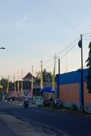 The video captures a street scene with a road, buildings, and several flags. The flags are predominantly yellow and yellow and blue, strung along poles and wires that crisscross the frame. A blue and orange building is visible on the right side of the road. There are also glimpses of people and motorcycles. The overall lighting suggests it is either dawn or dusk. The perspective is slightly tilted, making the scene appear sideways.