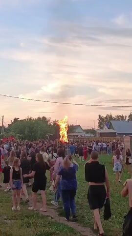 A large bonfire burns brightly in the center of the frame, its flames reaching high into the sky. A crowd of people, numbering in the hundreds, are gathered around the bonfire, some standing, some walking. The setting appears to be a rural area, with houses visible in the background and trees bordering the gathering. The sky is a mix of blue and soft clouds, suggesting either early morning or late afternoon. People of various ages are present, dressed in casual summer attire. The atmosphere appears festive and communal, with individuals interacting and moving throughout the space.