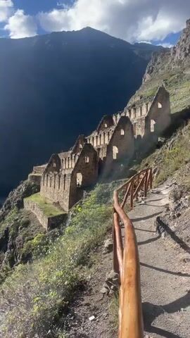 The video shows a path with a wooden handrail leading towards a stone structure with multiple rooms built into a mountainside. The structure has an old, weathered appearance, with open windows or doorways in each room. Behind the structure, a large mountain looms, partially shadowed. The sky is blue with scattered white clouds, and sunlight streams down, creating rays of light that illuminate the structure. The path is made of dirt and gravel, and some greenery grows along the mountainside. As the video pans up, a broader view of the valley below is revealed, showcasing agricultural fields and a small village nestled in the landscape. The sun is high in the sky, creating glare.