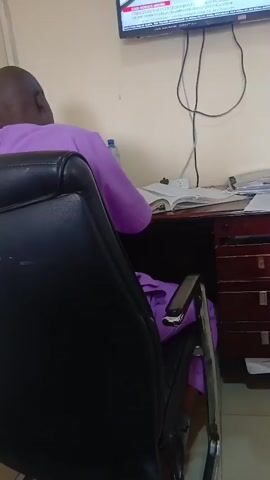 An adult male with a shaved head, wearing a light purple collared shirt, sits in a black office chair facing a desk. The desk has papers, an open book, and a water bottle on it. A dark brown file cabinet is built into the desk. A flat screen television is mounted on the wall above the desk, displaying news information. A power cord hangs from the television. The wall is painted a light neutral color.
