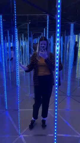 A woman films herself with a smartphone while standing in a mirror maze illuminated by blue LED lights. She wears a plaid shirt, a yellow t-shirt, black pants, and black and white sneakers. She is looking at the camera and occasionally interacts with a young boy in a white t-shirt and black sweatpants, who appears behind her in the maze. The camera is positioned in front of the woman, showing her reflection and the surrounding environment.