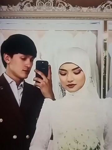 A young couple poses in front of a mirror, taking a selfie with a smartphone. The man wears a dark suit, a white shirt, and has short dark hair. The woman is dressed in a white hijab and gown. The background includes a decorative mirror frame and what appears to be wedding dresses on display. The lighting is bright, and there's a reflection of a ring light in the mirror. The woman briefly covers her mouth with her hand.