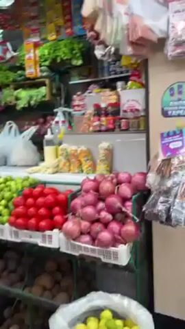 The camera pans across a bustling vegetable stand in a marketplace.  The stand is piled high with fresh produce including carrots, limes, tomatoes, onions, potatoes, and various greens.  Packaged goods are displayed on shelves behind the produce.  Several people are present, including customers shopping and vendors tending to the stand.  One customer wearing a black hoodie moves along the front of the stand.