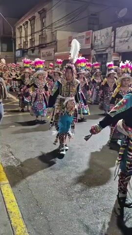 A group of people in colorful, ornate costumes are marching in a street at night. The costumes feature elaborate headdresses with feathers, colorful fabrics, and intricate embroidery. Some individuals are wearing masks or face paint. The street is paved and lined with buildings, some of which have signs. Streetlights illuminate the scene. The people appear to be participating in a parade or festival.