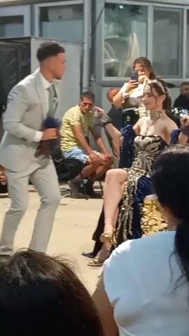 A man in a light gray suit walks towards a woman seated on a chair. The woman is wearing a formal, strapless, navy blue and gold embellished gown with her legs crossed. She has jewelry around her neck and her hair is styled up. She is holding a phone and appears to be taking a picture or recording. Another woman stands behind her, also holding a phone. In the background, there are other people seated and standing, and musical equipment is visible. The scene appears to be an outdoor event or gathering.