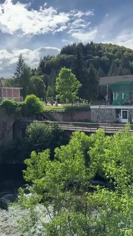 The camera films a stone bridge with an arch over a river in a green, outdoor setting. The sky is bright with moving clouds. Other buildings are visible, including a brick structure on the left, and a building with green siding on the right. Trees and bushes are prevalent throughout the landscape.  The river flows rapidly beneath the bridge. The camera moves slightly to the left, revealing more of the bridge's architecture.  No people are visible.