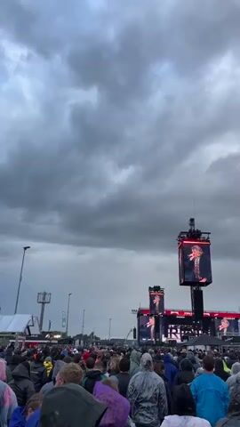 A large outdoor crowd, mostly facing a distant stage, stands under a heavily overcast sky. Many individuals in the foreground are wearing rain ponchos and jackets. The stage features multiple large LED screens displaying a male performer. Two tall towers, each topped with a large LED screen, are positioned further into the crowd. At the 0:01 mark, multiple columns of fire erupt from the stage, followed by a large, dark, spherical explosion of smoke high in the sky to the left of the main stage. The camera then pans upwards and right, briefly following the smoke before panning back down to the stage and the rightmost screen tower.