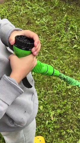 A close-up shot shows a child's hands holding a green garden hose with a black nozzle. The nozzle has multiple settings, including "JET" and "MIST," which are visible. The child appears to be turning the nozzle. The background is a grassy area. The child is wearing a gray sweatshirt and yellow Crocs.