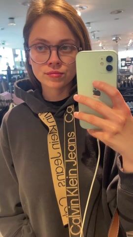 A young woman wearing glasses and a black Calvin Klein hoodie takes a selfie in a clothing store. She holds a light green iPhone in her right hand. The background shows clothing racks and other shoppers. The lighting is bright.