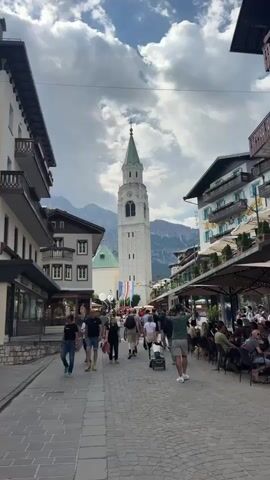 The video shows a wide cobblestone street lined with buildings. In the background, a tall, white church steeple with a green roof rises above the structures. Mountains are visible behind the buildings, with clouds in the sky. A crowd of people are walking down the street, some are sitting at outdoor cafes. There are umbrellas providing shade for the outdoor seating areas. The buildings on either side of the street have balconies and shops on the ground floor. The overall impression is a bustling European mountain town.