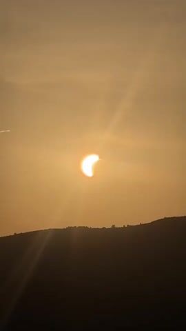 The video shows a partial solar eclipse taking place during sunset. The sun, partially obscured by the moon, is a bright, warm orange orb in a hazy, golden sky. The sun's rays are visible, fanning out from the celestial body. In the foreground, a silhouetted landscape of rolling hills stretches across the bottom of the frame, appearing dark against the illuminated sky. The faint outline of a crescent shape, possibly another part of the eclipse or a cloud formation, is visible higher in the sky. The overall impression is one of natural beauty and cosmic spectacle.