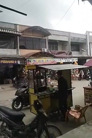 A street scene, likely in Southeast Asia, shows a man in a black shirt and black pants standing behind a yellow food cart. The food cart has a canopy and appears to be made of metal and wood. It is on wheels. In front of the cart, there is a black motorcycle. To the left of the cart is another black motorcycle. Behind the cart are several shops with awnings. One shop has a sign that reads "FAUZY PENANG 2". Another shop has a sign with colorful cartoon characters and the words "MUJUR JAYA". There are also stacks of what appear to be inflatable balls visible in front of the shops. The sky is overcast. The ground is paved. The man behind the cart is looking down and appears to be preparing food or interacting with a customer who is not fully visible.