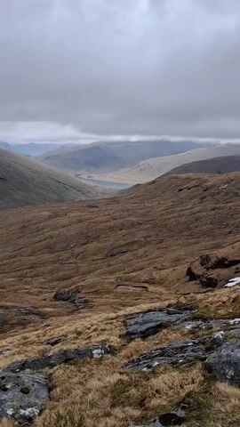 The video shows a landscape of mountainous terrain on a cloudy day. The mountains are primarily covered in brown and tan vegetation with some rocky outcroppings.  In the distance, a body of water can be seen nestled between the hills. The camera pans across the scene, revealing a faint path winding down one of the slopes. Towards the end of the video, two people are visible hiking along a rocky ridge. One is wearing a red jacket, and the other is carrying a white bag on their back. The overall environment appears rugged and somewhat desolate.