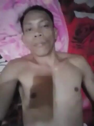 This is a low-resolution, blurry video showing a shirtless man lying down. The background appears to be a bed with a pink and red floral pattern. The man's face is partially visible and he is looking towards the camera. The video is very short and does not contain any discernible actions or interactions.