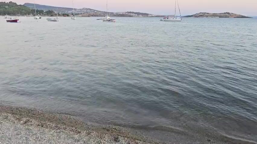 The video shows a tranquil ocean scene at dusk. Several sailboats are anchored or slowly moving in the water. The sky is a soft gradient of pink and blue, with a faint crescent moon visible. The foreground features a pebbly shoreline with gentle waves lapping at the edge. In the distance, a coastline with scattered buildings and trees can be seen. The overall atmosphere is peaceful and serene.