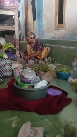 The video shows an elderly woman crouching down and interacting with items on a cluttered floor. The environment appears to be an indoor space, possibly a kitchen or storage area, with a tiled floor and unpainted walls. There are various objects scattered around, including plastic containers, bowls, bottles, and what appears to be fresh produce or herbs. A red cloth is draped over some of the items. The woman is wearing a patterned shirt and yellow pants. The lighting is dim.