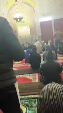 The video shows the interior of a mosque, where a group of people are kneeling on prayer rugs, facing forward.  They appear to be engaged in prayer or a religious ceremony.  The camera holder is standing at the back of the room, slightly to the side. The mosque has ornate decorations, including an archway with Arabic calligraphy.  A chandelier hangs from the ceiling.  Natural light comes through a doorway at the back of the room.  People are dressed in a variety of casual clothing, some wearing hoodies or jackets.
