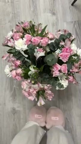 Here's a description of the video:

The video shows a large bouquet of pink and white flowers in a vase, set on a gray wood-patterned floor. The bouquet contains pink roses, white carnations, pink alstroemeria, and eucalyptus leaves. A person wearing white pants and pink slippers is standing behind the bouquet. A hand with pink painted nails reaches into the bouquet, gently touching and adjusting the flowers. A muffled voice can be heard in the background.