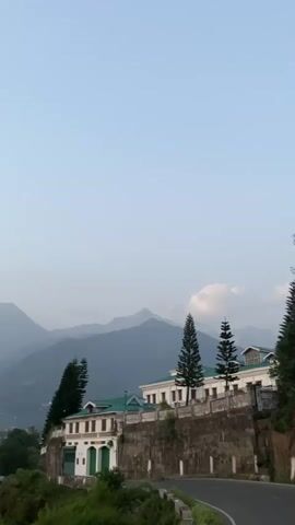 The video pans from right to left across a mountainous landscape. In the foreground, a white building with a green roof is perched on a stone wall overlooking a winding road. Several tall, slender pine trees stand beside the building. In the background, several layers of hazy, blue-gray mountains recede into the distance. The sky is a pale, clear blue with a few wispy clouds. The scene appears to be captured at dusk or dawn due to the soft, diffused light.