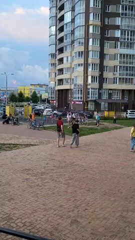 Two young males are walking in the foreground on a sandy area. In the background, a tall building with many windows is visible, along with a playground, parked cars, and other people. The sky is cloudy.