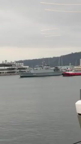 The video shows a gray warship labeled M270 docked in a harbor. On the warship, there are three flags. The colors on the top flag are not clear. The middle flag is white and red. The bottom flag is white, green, and red. Several people are visible on the warship. In the background is a harbor with other boats and buildings. The sky is cloudy. The video is taken from a boat looking out through a window with some light reflection on the window.