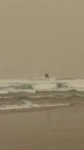 The video shows a kitesurfer in action on a stormy beach. The sand is wet, and the waves are moderately high. The sky is overcast, lending a muted, greyish tone to the scene. The kitesurfer is seen initially performing a jump, then landing back on the water, with the colorful kite clearly visible above.