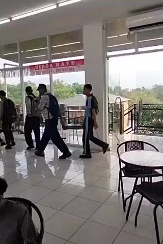 The video shows a group of young people, some carrying backpacks, walking into a brightly lit indoor space with large glass windows. The floor is tiled with a light-colored pattern. Outside the windows, there is greenery and some buildings. A banner with red and white lettering is visible through the windows, partially obscured by the frames. Some of the individuals are walking towards tables and chairs arranged in an outdoor seating area visible through the glass. In the foreground, two individuals are seated at a table.
