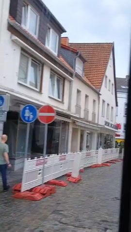 The video shows a view from inside a vehicle, likely a bus, looking out at a European street scene. The street is paved with cobblestones and lined with buildings. In the foreground, there are white construction barriers with orange bases, indicating ongoing work. The buildings vary in architectural style, with some featuring traditional half-timbered facades and others having more modern designs. Some shopfronts are visible, with signs and displays. The sky is overcast. The camera pans slowly, offering glimpses of different buildings and street details. There are no people clearly visible in the footage.