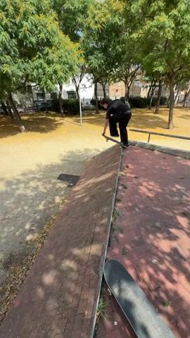 The media file shows a person skateboarding in an outdoor park setting. The person is wearing black pants, a black t-shirt, and white sneakers. They are seen performing a skateboarding trick, coming down a brick incline and then riding on a sandy path. Another skateboard is visible on the ground in the later frames. The scene is set on a sunny day, with dappled sunlight filtering through trees, creating shadows on the ground. Yellow leaves are scattered across the sandy area, suggesting it might be autumn. In the background, there are trees, buildings, and a fence. The overall atmosphere appears casual and recreational. The location is identified as Viladecans, Spain. No specific emotions are discernible, but the activity suggests a focus on skateboarding. There is visible text on the person's underwear, reading "Calvin Klein".