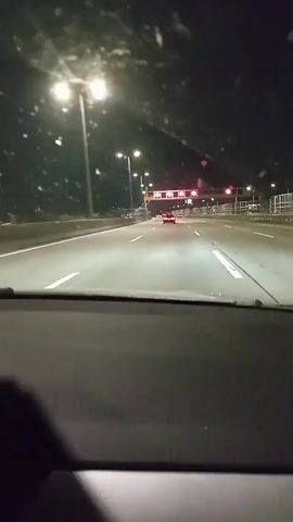 The video shows a first-person view from inside a vehicle driving on a highway at night. The dashboard is visible in the foreground. The road ahead is illuminated by streetlights and the headlights of other vehicles. There are multiple lanes of traffic, and several cars can be seen in the distance. Overhead signs display speed limits, with one sign clearly showing "80" in red circles. The windshield has numerous small white specks, possibly dirt or rain. The overall atmosphere is dark and the road appears wet.