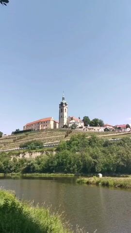 The video shows a wide shot of a river in the foreground and a castle or monastery on a hill in the background. The sky is clear and blue. The castle has a tall tower with a green roof and a spire. There are buildings with orange roofs next to the tower, and terraced vineyards on the hillside leading up to the castle. Trees and bushes are visible on the lower slopes of the hill and along the riverbank. The water in the river is calm and reflects the sky. There are no people or animals visible in the video. The camera appears to be stationary, capturing a landscape view.