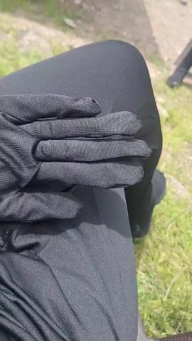 A person wearing black gloves and black leggings sits in an outdoor environment. The person's gloved hand is resting on their leg. Grass and a dirt path are visible in the background. The person moves their hand and opens it, then closes it.