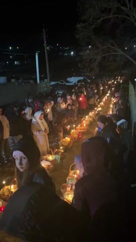 At night, a large group of people stand along a narrow walkway, each holding a candle and many with baskets. Some people are wearing winter clothes. The baskets are filled with what appear to be foods and decorations and also contain candles. The lit candles illuminate the people's faces. There are parked cars visible in the background, along with a streetlight. A tree is seen on the right side of the walkway. The air is still, and the overall ambiance is calm and reverent.