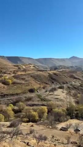 The video shows a landscape featuring dry, rolling hills under a clear blue sky. The sun is shining brightly, and in the valley below, a winding river flows through trees that are tinged with autumn colors. A narrow road cuts through the scene, adding a human element to the otherwise natural setting. The overall impression is one of serene beauty and tranquility.