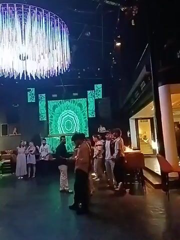 The video depicts a contemporary venue, possibly a lounge or event space, with a diverse crowd milling around. A large, round, multi-tiered LED chandelier dominates the ceiling, emitting a bright, multicolored light that adds to the ambient atmosphere. A sizable LED screen in the background displays kaleidoscopic patterns that morph in color and design, flanked by smaller screens presenting similar abstract visuals.

People of different ages and styles are visible, some engaged in conversation, while others stand in small groups, apparently waiting. The clothing worn ranges from casual to semi-formal, indicating a relaxed yet upscale setting.

The camera pans across the scene, offering a wide perspective of the space, which seems to be set up for an event or gathering, though the specific purpose remains unclear from the available footage.