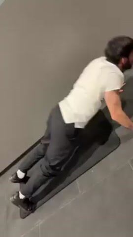 A man is performing wall push-ups. He is wearing black shoes with white socks, black pants, and a white t-shirt. He is using a black yoga mat. The wall is gray, and the floor is gray tiles.