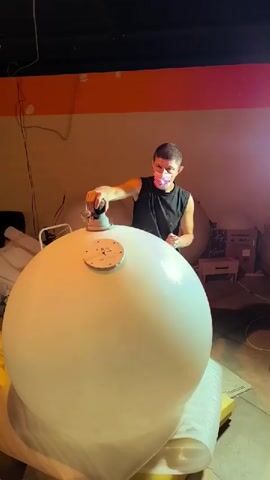 A man wearing a black tank top and a pink face mask is sanding the top of a large, white, spherical object. The object appears to be made of a smooth, possibly plastic material, and is resting on a surface covered with white protective material. The man is holding a sanding tool with both hands and applying it to a circular wooden plate attached to the top of the sphere, which has several screws visible. The background is dimly lit, with a yellow-orange stripe visible on the wall. Other spheres of similar size and color are visible in the background. The scene appears to be an indoor workshop or storage area.
