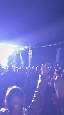 A video shows a lively rock concert from the perspective of an audience member in the foreground. The stage, illuminated by bright lights, features a band performing, with a large screen behind them displaying the words "РАБОТНИКИ КУЛЬТУРЫ" (Workers of Culture). The audience, a mix of men and women, is actively engaged, dancing, swaying, and recording the performance on their phones. The atmosphere is energetic, filled with the sounds of live music and cheers. The overall scene is vibrant and dynamic, capturing the excitement of a concert.