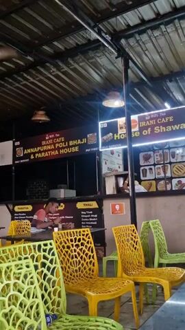 The video shows an outdoor seating area of a food stall with a corrugated metal roof. In the background, there are signs for "IRANI CAFE" and "House of Shawarma" with pictures of food items. A person wearing a red shirt is sitting at a table to the left, eating. Brightly colored plastic chairs, yellow and green, are arranged around tables in the foreground. The lighting is dim, with overhead lamps illuminating the area.