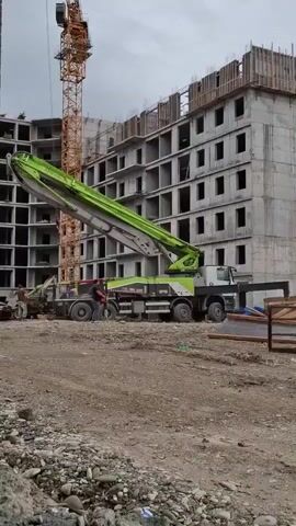 A concrete pump truck is positioned on a construction site. The truck is light-colored with green detailing on its extended boom. Two individuals are visible near the base of the boom, one of whom appears to be interacting with the machinery. A tall yellow tower crane stands to the left of the truck, and a multi-story building under construction occupies the background. The ground is covered in gravel and dirt. The sky is overcast.