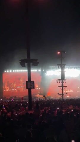 The video captures an expansive night-time view of a large outdoor music festival or concert in Indio, United States of America. A vast crowd of attendees fills the scene, stretching from the foreground into the midground, with many individuals holding up their cell phones to record the event. In the background, a brightly illuminated stage is visible, featuring multiple large video screens displaying dynamic visuals that frequently shift between reddish-orange and blue hues. The stage is equipped with numerous powerful lights, casting beams through the smoke or haze rising from the stage area.

Two prominent, tall light towers stand within the crowd. The one in the center-left displays a "D5" sign, likely indicating a section or zone. Both towers are outfitted with multiple spotlights pointing towards the stage and audience. The overall atmosphere appears energetic and engaging, typical of a live music event. A small flag, exhibiting green, white, and red vertical stripes, possibly the flag of Mexico, can be seen within the crowd briefly.