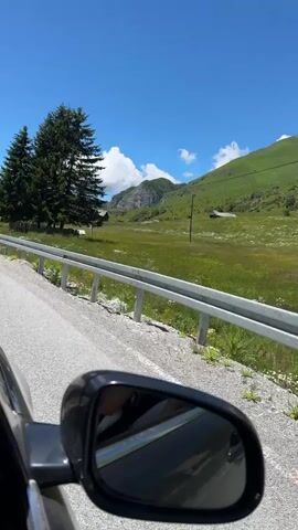 The video captures a scenic view from a car, looking out towards a lush green landscape under a clear blue sky with scattered clouds. In the foreground, a car's side mirror reflects the interior of the car and the road ahead. Beyond the mirror, a guardrail separates the road from a grassy field dotted with wildflowers. Several tall evergreen trees stand prominently in the middle ground, with some buildings visible behind them. The terrain rises into rolling green hills and mountains in the distance. The perspective suggests the car is in motion, driving along a paved road that curves to the left.