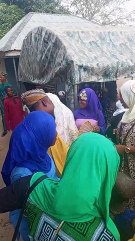The video shows a group of people, mostly women wearing hijabs, gathered outdoors. The women are dressed in brightly colored clothing and appear to be celebrating or socializing. One woman in a purple hijab holds a decorative fan. Some of the women are laughing and talking. A man in a yellow shirt and patterned hat interacts with the group. In the background, there's a makeshift structure with a corrugated metal roof and fabric draped over it. The environment looks like a village setting, with trees and simple buildings nearby.