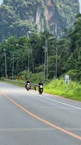 Two individuals on motorcycles are traveling down a paved road. One motorcycle, on the left side of the frame, is pulling a small, red, single-wheel trailer. The other motorcycle, on the right, is traveling slightly behind and to the right of the first. Both motorcycles appear to be moving in the same direction. The road is flanked by lush green vegetation and palm trees. In the background, a large, steep, green, mountainous landscape dominates the scene. The sky is bright, suggesting daytime. There are utility poles and wires running along the side of the road. A white sign with the number 8 is visible on the right side of the road. The road has a double yellow line in the center and a single yellow line on the left edge. Yellow curve warning signs are visible on the left side of the road.