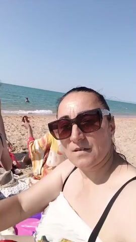 The video begins with a close-up of a woman wearing sunglasses and a white top, sitting on a beach towel. She is looking directly at the camera. The background shows a sandy beach, the ocean, and a clear blue sky. A person in a bikini with red and white striped straps is sitting on the sand, facing away from the camera, with their back to the viewer. Behind them, another person in a black bikini is standing, holding a bottle of soda and a white towel, and appears to be handing something to the person sitting. Further in the background, other people can be seen near the water. The video pans slightly to the left, revealing more of the beach and the sea.