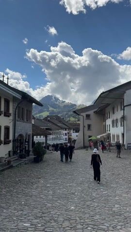 The video shows a cobblestone street in a quaint European village. Several people are walking down the street, including a woman in a leopard print dress who walks past the camera. The buildings on either side of the street are made of stone and have a traditional European architectural style. In the background, a large mountain with snow-capped peaks can be seen. The sky is blue with fluffy white clouds, suggesting a pleasant day. The video appears to be taken at ground level, giving the viewer a pedestrian's perspective of the street scene.